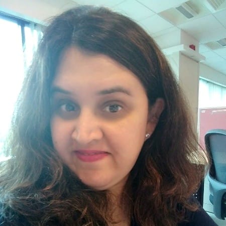 A woman with wavy brown hair and light skin, wearing a dark top, smiles slightly while sitting in an office with large windows and modern furniture in the background.