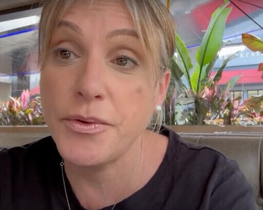 Alison Nightingale, a woman with light hair and pearl earrings, sits indoors near a window with plants outside. She is wearing a black top and looking slightly to the side.
